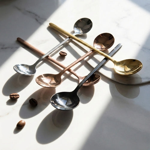 Artistic minimalist shot of the four-piece spoon set (gold, silver, black, rose gold) on white marble, emphasizing dramatic shadows and metallic reflections.
