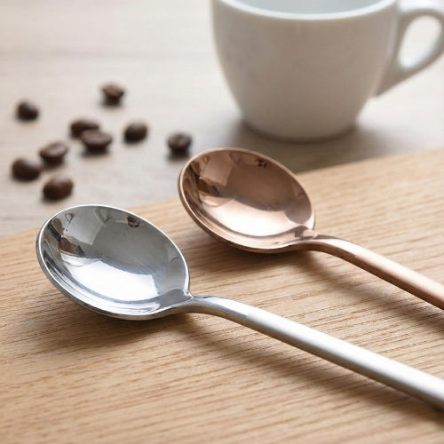 Hand stirring espresso with a gold spoon, featuring a blurred premium stainless steel French Press and croissant in the background for an elegant breakfast vibe.