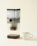Clear plastic cold brew coffee maker with ice and coffee grounds, next to a white cup