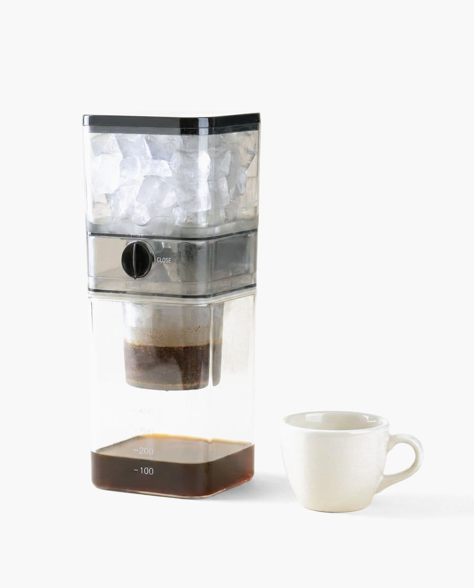 Clear plastic cold brew coffee maker with ice and coffee grounds, next to a white cup